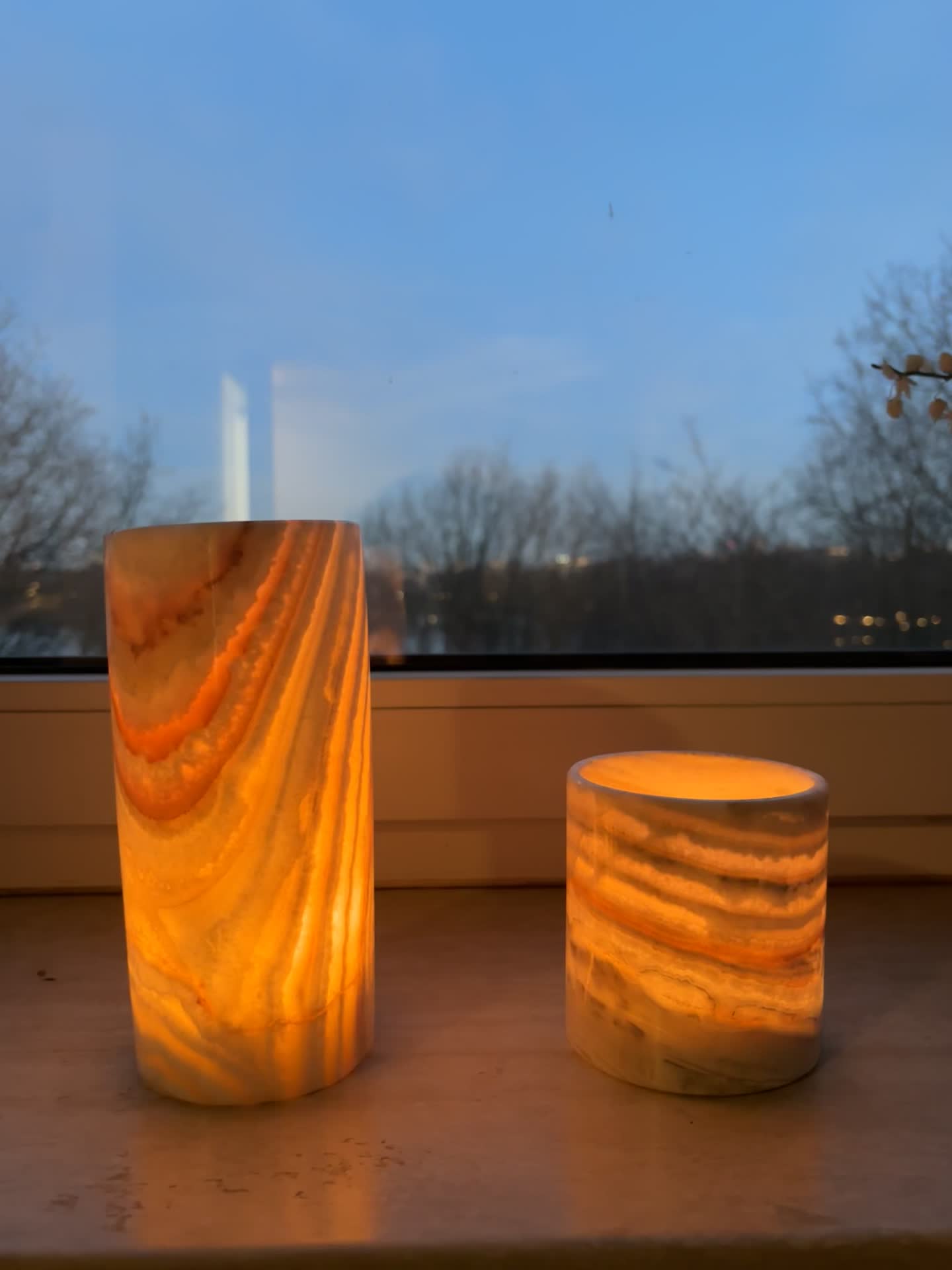 orange-washed cylindrical alabaster candle holders on a reflective surface with a window view.