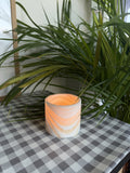 The Vessel S, a straight-sided polished alabaster tealight cup glowing warm, on a checked cloth beside palm leaves