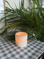 The Vessel S, a straight-sided polished alabaster tealight cup glowing warm, on a checked cloth beside palm leaves