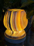 The Barrel alabaster lamp glowing amber, its rounded symmetric form banded in orange and grey on a ridged wood base