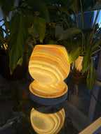 The Boulder lit, a rounded turned alabaster lamp glowing amber with concentric ring banding, on an alabaster collar and ridged wood base