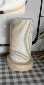 The Pillar L unlit in daylight, a tall alabaster column with olive and cream wavy banding on a turned wood base