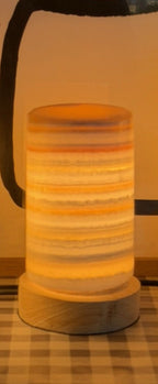 The Pillar M lit on its own, warm amber glowing through fine horizontal cream and rust banding, on a turned wood base