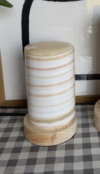 The Pillar M unlit in daylight, a compact alabaster cylinder with fine orange horizontal stripes on a pale wood base