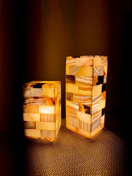 Mosaic Cube Pair lit: two tiled alabaster block lamps, one medium and one taller large, glowing amber on a woven mat in a dark room