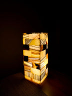 Mosaic Cube L lit alone in a dark room, a tall column of amber, grey and cream alabaster tiles with black tiles and a brown fossil-like inclusion