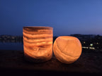The Duo lit on a stone ledge at dusk, banded cylinder and smaller orb glowing amber against water and distant lights